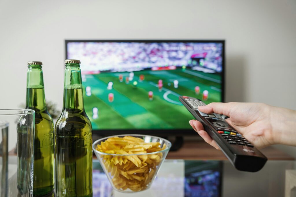 A person holding a remote while watching soccer on TV with beer and snacks on the table. Smart TV
